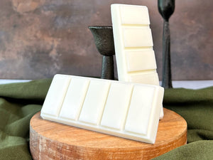 White wax melt bars stacked on a wooden block with a green fabric and dark candle holder in the background.