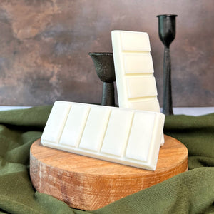 White wax melt bars stacked on a wooden block with a green fabric and dark candle holder in the background.