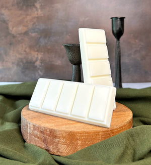 White wax melt bars stacked on a wooden block with a green fabric and dark candle holder in the background.