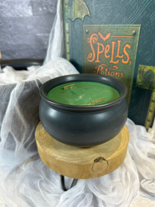 Photo showing 20 oz size Voo-Dew Candle. The candle is in a   black cauldron-shaped soup bowl. Surrounding the candles is a large black cauldron, a spell book, and spooky cheesecloth.