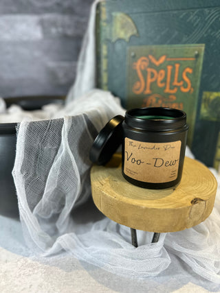 Photo showing 9 oz size Voo-Dew Candle. The candle tin is black with a metal lid. The candle description tag is brown colored. Surrounding the candles is a large black cauldron, a spell book, and spooky cheesecloth. 