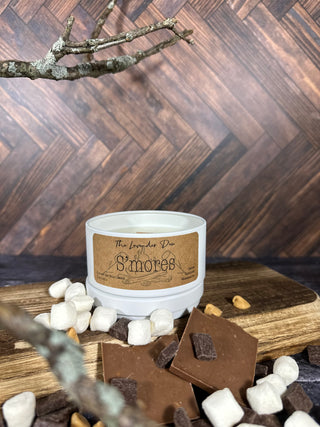 Photo showing 5 oz size S'mores Candle. The candle tin is a cream color with a metal lid. The candle description tag is brown. Surrounding the candle is wood, chocolate chunks, and marshmallows.