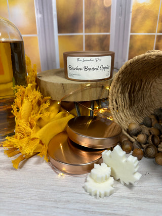 Photo showing 5 oz size Bourbon Braised Apples Candle. The candle tin is a copper color with a metal lid. The candle description tag is ivory colored. Surrounding the candle is a bottle of bourbon and acorns.
