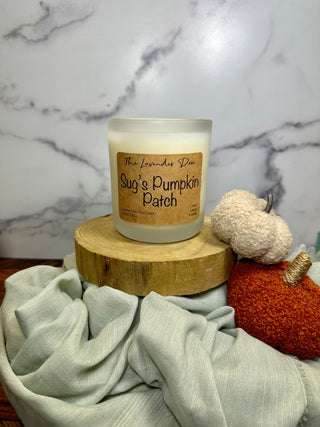 Photo showing 12 oz size Sug's Pumpkin Patch Candle. The candle jar is frosted glass with a brown-colored description tag. Surrounding the candle are fabric pumpkins, pale blue fabric, and wood accents.