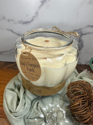 Photo showing 50 oz size Sug's Pumpkin Patch Candle. The candle is in a  glass pumpkin-shaped bowl with a matching lid. Surrounding the candle is pale blue fabric and wood accents.