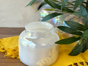 An iridescent  clear glass jar candle with white wax sits on a yellow fringed cloth on a wooden surface, with green leaves and a glass orb in the background.