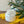 An iridescent  clear glass jar candle with white wax sits on a yellow fringed cloth on a wooden surface, with green leaves and a glass orb in the background.