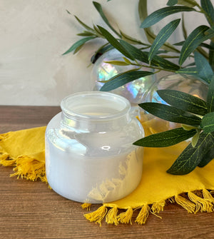 An iridescent  clear glass jar candle with white wax sits on a yellow fringed cloth on a wooden surface, with green leaves and a glass orb in the background.