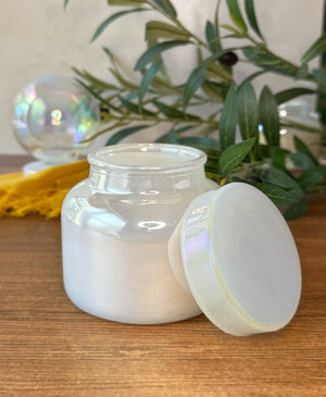 An iridescent glass jar filled with wax, shown with its matching lid leaning against it on a wooden surface. Green leaves and a yellow fabric are visible in the softly blurred background.