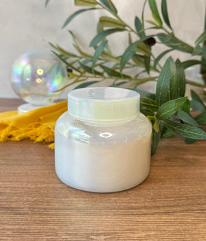 An iridescent jar containing wax with a match lid on top of the jar. The jar sits on a wooden surface with yellow fringed fabric, greenery, and a glass orb visible in the softly blurred background