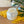 An iridescent glass jar filled with white wax, placed on a yellow fringed cloth on a wooden surface, with green leaves and glass orbs in the background.