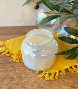 An iridescent glass jar filled with white wax, placed on a yellow fringed cloth on a wooden surface, with green leaves and glass orbs in the background.