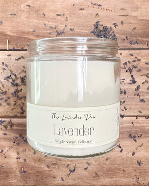 Lavender candle in a glass jar on a wooden surface with scattered lavender flowers.