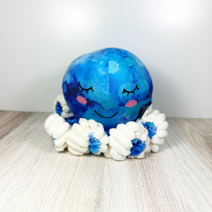 Plush blue tie-dyed octopus dog toy with white accents and a smiling face. Toy is sitting on a light wooden surface with a white background.
