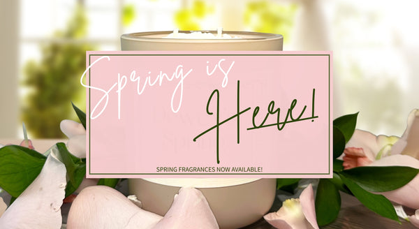 Spring candle container on a wooden surface surrounded by flower petals and greenery. Overlaying the image is a pink text block saying 'Spring is Here! Spring fragrances now available!'.