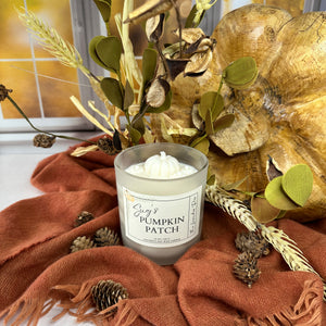 Candle labeled 'Sug's Pumpkin Patch' with decorative fall elements including a gold leaf, green leaves, and a orange fabric.