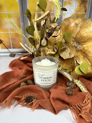 Candle labeled 'Sug's Pumpkin Patch' with decorative fall elements including a gold leaf, green leaves, and a orange fabric.
