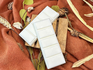 Two white wax melt bars on a wooden block with autumn-themed decor including leaves and pinecones on an orange fabric background.