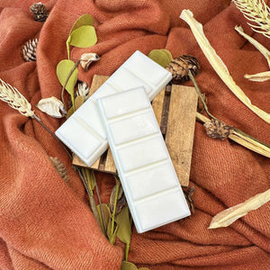 Two white wax melt bars on a wooden block with autumn-themed decor including leaves and pinecones on an orange fabric background.