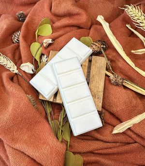 Two white wax melt bars on a wooden block with autumn-themed decor including leaves and pinecones on an orange fabric background.