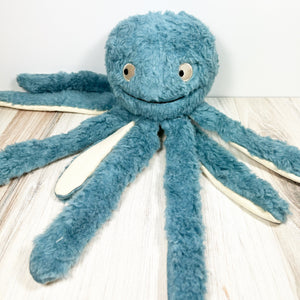 Blue plush dog toy resembling an octopus with long tentacles. The toy sits on a light wooden surface with a white background. 