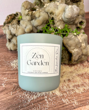 Candle with scent label 'Zen Garden' on a wooden surface surrounded by decorative rocks, plants, and a sprinkling of sand.